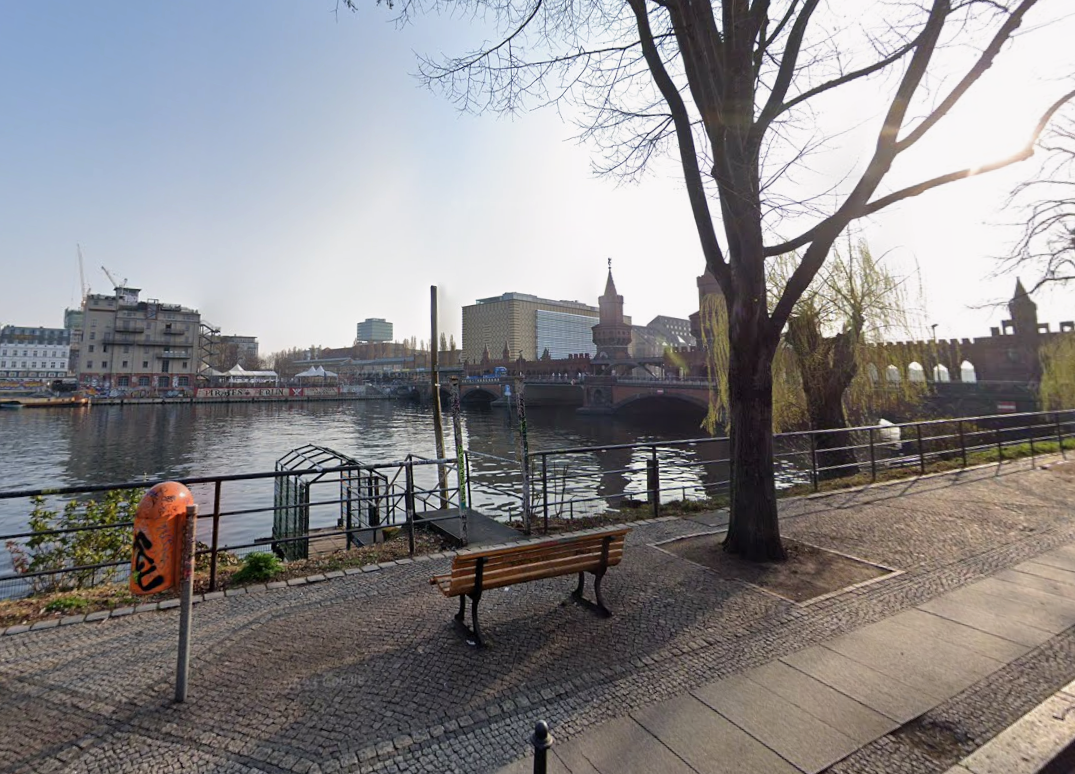 The Spree riverbank at Schlesisches Tor, looking toward the Oberbaumbrücke. A bench and railing along a stone path right by the water.